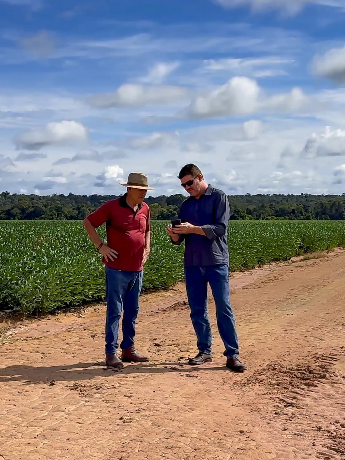 Um produtor e um homem mais jovem na estrada de terra ao lado da lavoura de soja, conferindo números no celular